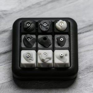 May include: A square mechanical keyboard macro pad featuring nine individual keycaps arranged in a three-by-three grid. Each keycap is sculpted to resemble a rock climbing hold in various shades of black, grey, and white, with a metal bolt.