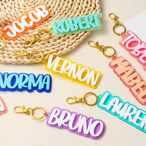Op de afbeelding: Een collectie kleurrijke acryl naamsleutelhangers met goudkleurige metalen karabijnsluitingen en ringen. Elke sleutelhanger toont een naam in witte blokletters op een transparante gekleurde acrylbasis, waaronder namen zoals Jacob, Robert, Tobey, Mabel, Vernon, Norma, Lauren en Bruno.