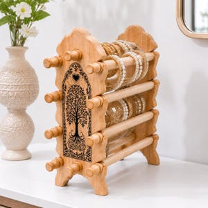 May include: A wooden bracelet display stand with a tree of life design. The stand is made of light-colored wood and features multiple rods for displaying bracelets. The tree of life design is black and is centered on the front of the stand. The stand is filled with various bracelets.