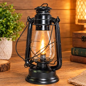 Può includere: Una lanterna da tempesta in metallo nero con un cilindro in vetro trasparente e una gabbia in filo metallico. La lanterna presenta una base circolare, una maniglia superiore e una fonte luminosa centrale che emette un effetto fiamma calda.