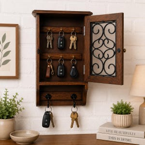 Pode incluir: Um porta-chaves de madeira castanha escura com uma porta articulada e trabalhos em ferro decorativos. O porta-chaves tem vários ganchos para pendurar chaves. A porta está aberta, revelando chaves em ganchos. O porta-chaves está montado numa parede de tijolo branco.