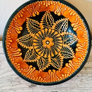 May include: A round ceramic bowl featuring a black center with a prominent orange and yellow floral mandala design. The wide rim is decorated with a repeating orange and yellow pattern of curved lines and dots against a dark background.