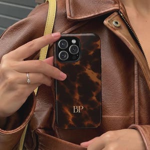 Pode incluir: Uma capa de smartphone com um padrão de tartaruga em castanho escuro e preto, com as iniciais douradas BP impressas no centro inferior. A capa tem um recorte para um sistema de câmara de lente tripla e é segurada contra um casaco de pele.