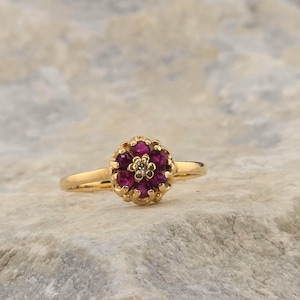 May include: A gold ring featuring a floral-shaped cluster setting. The center of the flower is a small, clear, round-cut stone surrounded by six vibrant, deep pink, round-cut stones. The band is a smooth, polished gold metal with a simple design.