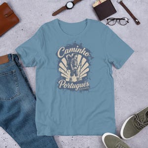 May include: A dusty blue t-shirt featuring a graphic print of a hiker with a backpack and walking stick centered over a scallop shell design. The text reads Caminho Portugu&ecirc;s and Bom Caminho in a vintage-style font with a distressed texture.