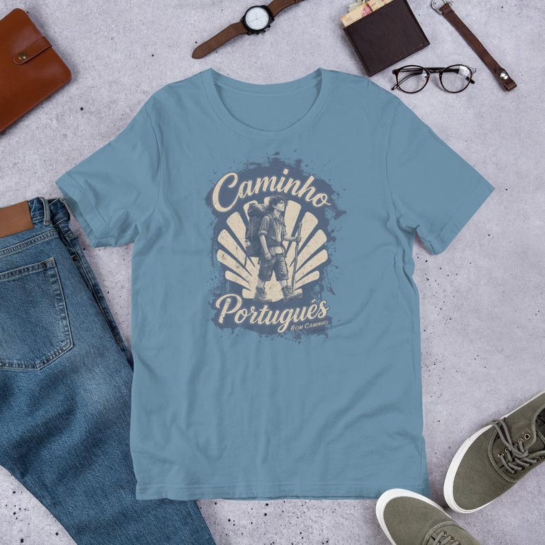 May include: A dusty blue t-shirt featuring a graphic print of a hiker with a backpack and walking stick centered over a scallop shell design. The text reads Caminho Portugu&ecirc;s and Bom Caminho in a vintage-style font with a distressed texture.