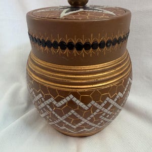 May include: A brown lidded jar featuring a geometric mosaic pattern in white and gold. The lid has a central knob and a star design. A horizontal band of black circular accents encircles the upper portion, while gold horizontal rings decorate the middle section.