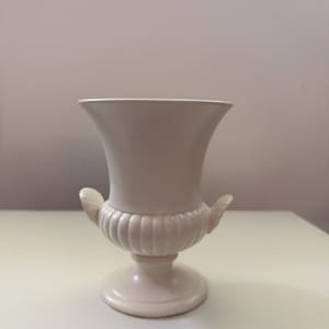 May include: A cream-colored urn-shaped vase with a flared top and a pedestal base. The vase features ribbed detailing around the midsection and decorative handles. The vase is approximately 7 inches tall.