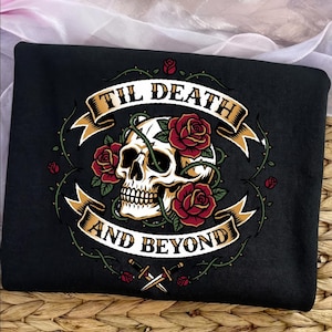 Puede incluir: Una camiseta negra con un estampado gráfico de un cráneo humano rodeado de rosas rojas y enredaderas verdes espinosas. Una cinta beige envuelve el diseño con el texto negro 'TIL DEATH AND BEYOND'. Dos dagas cruzadas se sitúan en la parte inferior.