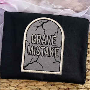 May include: A black t-shirt featuring a large embroidered patch shaped like a gray tombstone with black cracked detailing. The text GRAVE MISTAKE is stitched in black capital letters across the center of the tombstone, which has a thick white border.
