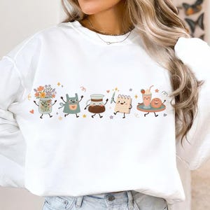 May include: A white long-sleeve sweatshirt featuring a horizontal graphic print of anthropomorphic items including a flower-filled mason jar, a teal apron, a coffee carafe, a notepad with a pencil, and a milkshake with a donut on a tray.