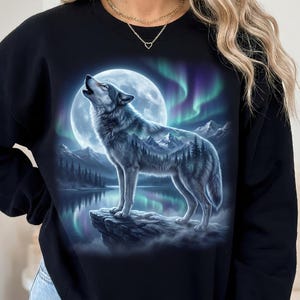 May include: A black crewneck sweatshirt featuring a graphic print of a wolf howling at a full moon. The wolf's body contains a double exposure landscape of mountains, pine trees, and a lake, set against a backdrop of vibrant green and purple aurora.