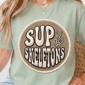 May include: A light sage green t-shirt featuring a circular patch with a tan background. The patch displays the text "SUP SKELETONS" in a bold, wavy, cream-colored font, accompanied by a graphic of a skeleton hand making a peace sign.