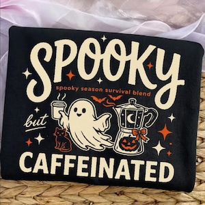 May include: A black t-shirt featuring a cream-colored graphic with the text SPOOKY but CAFFEINATED and spooky season survival blend. The design includes a ghost holding a coffee cup, a moka pot with a pumpkin, a black cat, bats, and stars.