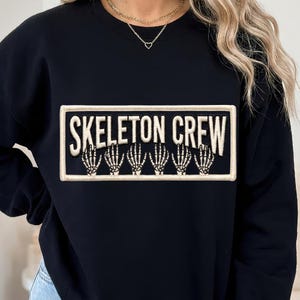 May include: A black crewneck sweatshirt featuring a rectangular patch with a cream-colored border. The patch displays the text SKELETON CREW in cream-colored lettering above a graphic of six skeletal hands arranged in a row on a black background.