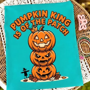 Puede incluir: Una camiseta de color verde azulado brillante con un gráfico de tres calabazas apiladas, la superior con una pequeña corona. El texto PUMPKIN KING IS OF THE PATCH aparece en letras naranjas. A la izquierda hay una silueta negra de un espantapájaros.