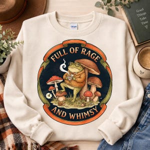 May include: A cream-colored crewneck sweatshirt featuring a graphic of a frog wearing glasses and a cardigan, holding a steaming cup. The frog is surrounded by mushrooms, a snail, and an acorn, with the text FULL OF RAGE AND WHIMSY.