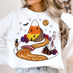 Op de afbeelding: Een wit sweatshirt met lange mouwen en een grafische print van een huis in de vorm van een candy corn met een trap, een kronkelend pad, een peperkoekhuisje, een zwarte kat, een vleermuis, sterren en een maan in oranje, geel, wit en bruin.