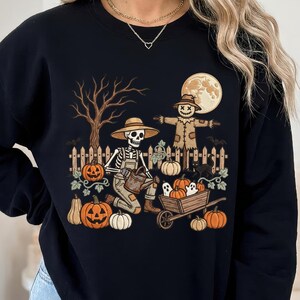 May include: A black long-sleeve sweatshirt featuring a graphic print of a skeleton wearing a straw hat and overalls, kneeling in a garden with a watering can. The scene includes a scarecrow, pumpkins, a wheelbarrow, ghosts, and a full moon.