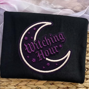 May include: A black sweatshirt featuring a large crescent moon patch in dark purple with a cream-colored embroidered outline. The text "Witching Hour" is embroidered in a gothic purple font across the center of the moon, accented by small purple star-shaped embroidery details.