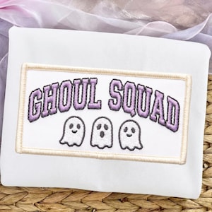 Peut inclure: Un sweat-shirt blanc avec un écusson rectangulaire brodé à bordure beige clair. L'écusson présente le texte GHOUL SQUAD en lettres violettes arquées au-dessus de trois petits fantômes blancs de style dessin animé aux contours noirs.