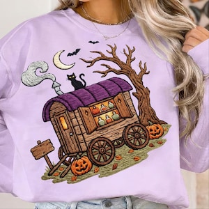 May include: A light purple sweatshirt featuring a graphic print of a wooden wagon with a purple roof, a black cat, a crescent moon, bats, a bare tree, and pumpkins. The wagon displays shelves filled with small colorful potion bottles.