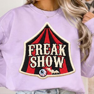 May include: A light purple crewneck sweatshirt featuring a large embroidered patch shaped like a circus tent. The patch has red and black vertical stripes, the text "FREAK SHOW" in cream, a single blue eyeball, and a small white bat silhouette.