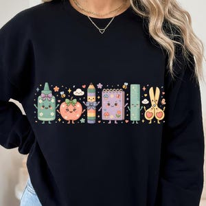 May include: A black crewneck sweatshirt featuring a colourful graphic print of anthropomorphic school supplies including a glue bottle, an apple, a pencil, a spiral notebook, a ruler, and scissors, all decorated with small flowers, hearts, and stars.