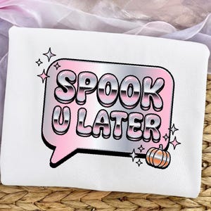 May include: A white sweatshirt featuring a graphic of a speech bubble with a pink and silver metallic gradient. The text reads SPOOK U LATER in bold, bubble-style letters. A small orange and silver pumpkin and several four-pointed star icons are included.