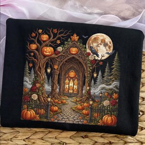 May include: A black sweatshirt featuring a detailed graphic of a gothic archway surrounded by autumn trees, pumpkins, and roses. A large full moon with a raven sits above the scene, while a stone path leads through the archway filled with glowing jack-o'-lanterns.
