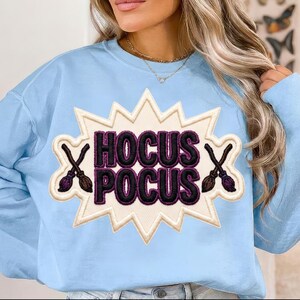May include: A light blue crewneck sweatshirt featuring a large, cream-colored, starburst-shaped patch. The patch displays the text HOCUS POCUS in bold, dark purple, embroidered letters, flanked by two small, crossed brown broomstick icons.