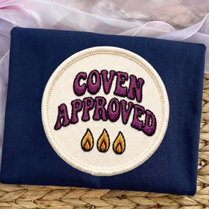 May include: A dark navy blue t-shirt featuring a circular embroidered patch. The patch has a cream-colored background with the purple embroidered text COVEN APPROVED and three small orange and yellow flame icons centered below the text.