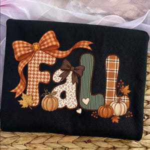 May include: A black sweatshirt featuring the word Fall in large, decorative letters. The letters are filled with various patterns including houndstooth, floral, and plaid. The design includes orange bows, small pumpkins, and autumn leaves.