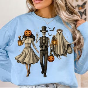 Könnte beinhalten: Ein hellblaues Sweatshirt mit einer grafischen Illustration von drei Figuren: eine Person mit Kürbiskopf, die eine Laterne hält, ein Skelett mit Zylinder und Weste, das einen Kürbiseimer trägt, und ein Geist mit Blumenkranz, der eine Laterne hält.