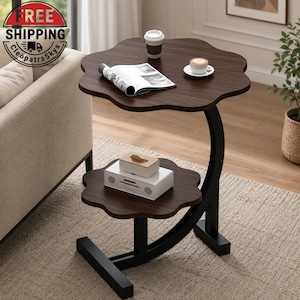 May include: A two-tier side table with a dark wood grain finish and a flower-shaped top and bottom shelf. The table features a black metal C-shaped frame. On the top surface sits an open magazine, a tall cup, and a small cup on a saucer.