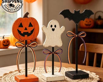 May include: Three Halloween tabletop decorations on black metal stands with square bases. One is an orange jack-o'-lantern, one is a white ghost, and one is a black bat. Each figure features a decorative bow made of orange, black, and white ribbon.