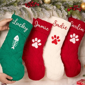 May include: Four knit Christmas stockings featuring ribbed cuffs and cable knit patterns. The green stocking displays a white fish skeleton, while the red and white stockings feature white or red paw prints. Each cuff is embroidered with names.