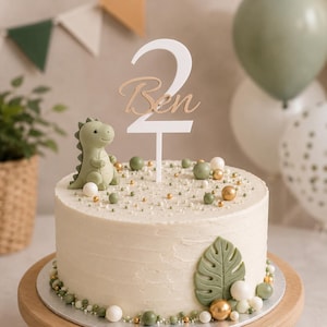 Könnte beinhalten: Ein runder Kuchen mit cremefarbener Glasur, verziert mit einer kleinen grünen Dinosaurierfigur, einem weiß-goldenen Tortenstecker mit der Zahl 2 und dem Namen Ben sowie einem grünen Blatt. Der Kuchen ist mit kleinen grünen und goldenen Kugeln belegt.