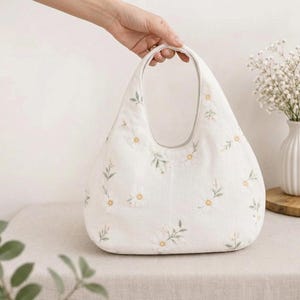 May include: A white hobo-style handbag featuring an embroidered pattern of small white daisies with yellow centres and green stems. The bag has a rounded silhouette with an integrated handle opening at the top, crafted from a textured fabric material.