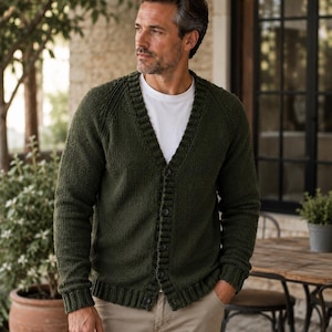 May include: A dark olive green knit cardigan featuring a V-neckline, long sleeves with ribbed cuffs, and a button-down front closure. The garment is worn over a plain white crew neck t-shirt and paired with light tan trousers.