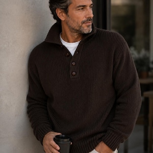 May include: A dark brown, long-sleeved, ribbed knit sweater featuring a high collar and a partial button-down placket with three dark brown buttons. The sweater is worn over a plain white t-shirt, paired with light-colored pants.