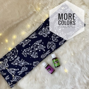 Può includere: Un impacco per il collo per aromaterapia blu navy con un motivo di costellazioni bianche. L'immagine mostra anche due flaconi di oli essenziali e il testo "MORE COLORS AVAILABLE". L'impacco è su una superficie bianca e soffice.