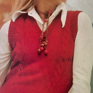 Może przedstawiać: Czerwony wełniany bezrękawnik z warkoczowym splotem, pod spodem biała koszula z kołnierzykiem. Bezrękawnik ma dekolt w kształcie litery V i brązowy skórzany sznurek z czerwonymi koralikami, który zwisa.