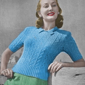 May include: A blue knitted sweater with a cable knit pattern and short sleeves. The sweater has a collared neckline and is worn by a woman with red lipstick.
