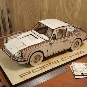 Puede incluir: Maqueta de madera de un coche deportivo Porsche clásico, con una intrincada construcción en capas. El coche es de color claro con detalles en marrón oscuro, y se encuentra sobre una base de madera con el logotipo "PORSCHE". El modelo incluye ruedas, ventanas e interior.