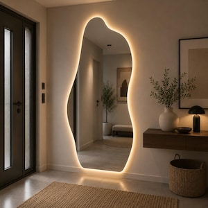 Puede incluir: Un espejo de pared de cuerpo entero con forma orgánica irregular y retroiluminación LED cálida integrada. El espejo está montado en una pared lisa y refleja el interior de un pasillo con una planta en maceta, un banco y una puerta al fondo.