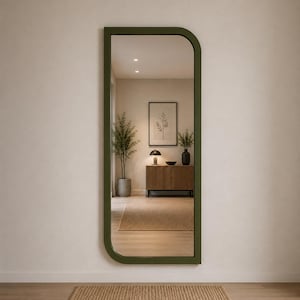 Op de afbeelding: Een hoge, rechthoekige wandspiegel met een donker olijfgroen frame, voorzien van een afgeronde rechterbovenhoek en een vierkante linkerbenedenhoek. Het spiegeloppervlak toont een kamerinterieur met een houten kast, een lamp en een plant.