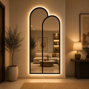 May include: A tall, floor-standing mirror featuring two overlapping arched frames with a black metal border. The mirror is illuminated by integrated LED backlighting that casts a warm glow against the wall, creating a distinct silhouette.