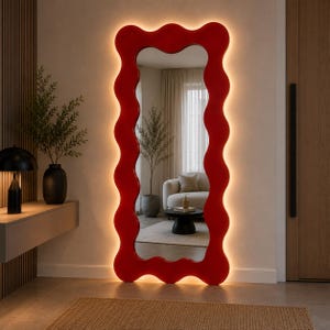 May include: A full-length floor mirror featuring a thick, wavy, red frame. The mirror is illuminated from behind by a warm, glowing LED light strip that casts a soft light onto the wall, highlighting the undulating shape of the frame.