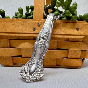 May include: A silver-colored spoon handle repurposed as a keychain. The handle features an ornate grape and leaf design. The keychain is attached to a silver split ring. The background includes a wooden basket and green plant.